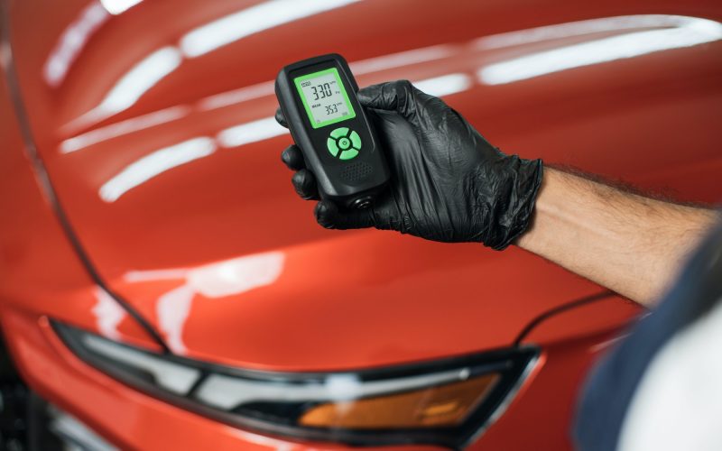 Closeup view of male hand, using film coating thickness gauge device to check car paint.